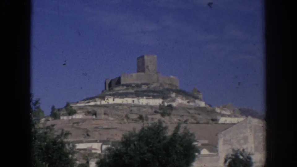 1966 Spain 8mm frame showing a hilltop stone castle above whitewashed buildings, trees, and blue sky.