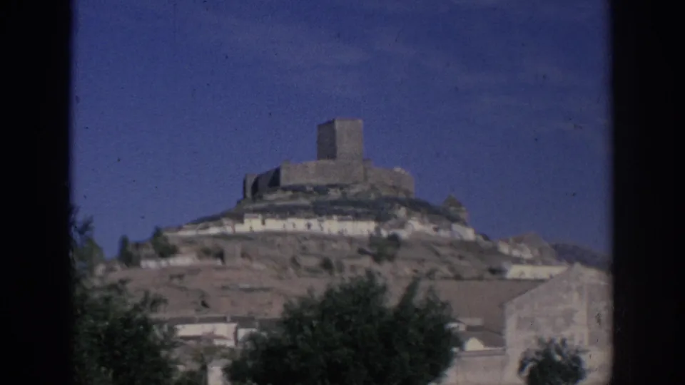 1966 Spain home-movie frame with a hilltop fortress, pale buildings at its base, trees, and a clear blue sky.