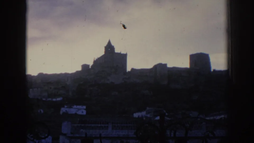 1966 Spain home-movie frame with a silhouetted castle skyline, tower forms, dark foreground buildings, and muted sky.