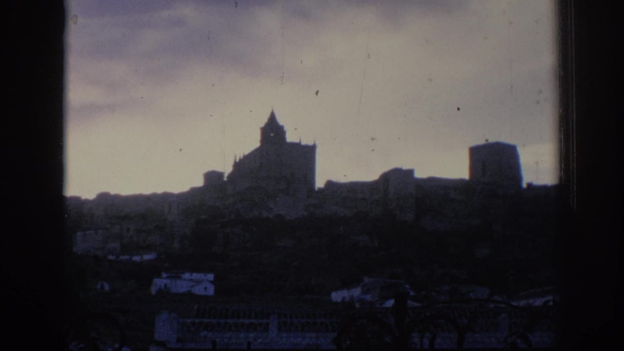 1966 Spain home-movie frame looking through a dark opening toward a silhouetted castle-like structure on a hill above low buildings.
