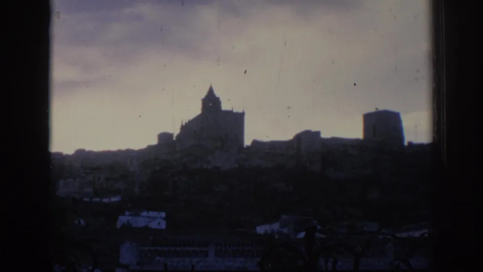 1966 Spain home-movie frame looking through a dark opening toward a silhouetted castle-like structure on a hill above low buildings.