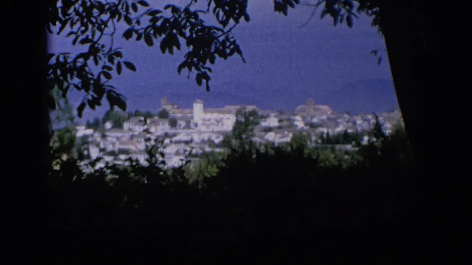 1966 Spain travel footage shows a distant white town and bright tower viewed from beneath trees against a blue backdrop.