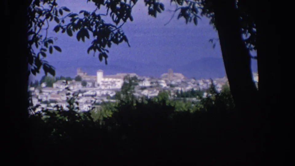 1966 Spain home-movie still frames a distant hillside or coastal town through dark trees, with white buildings and a tower visible.
