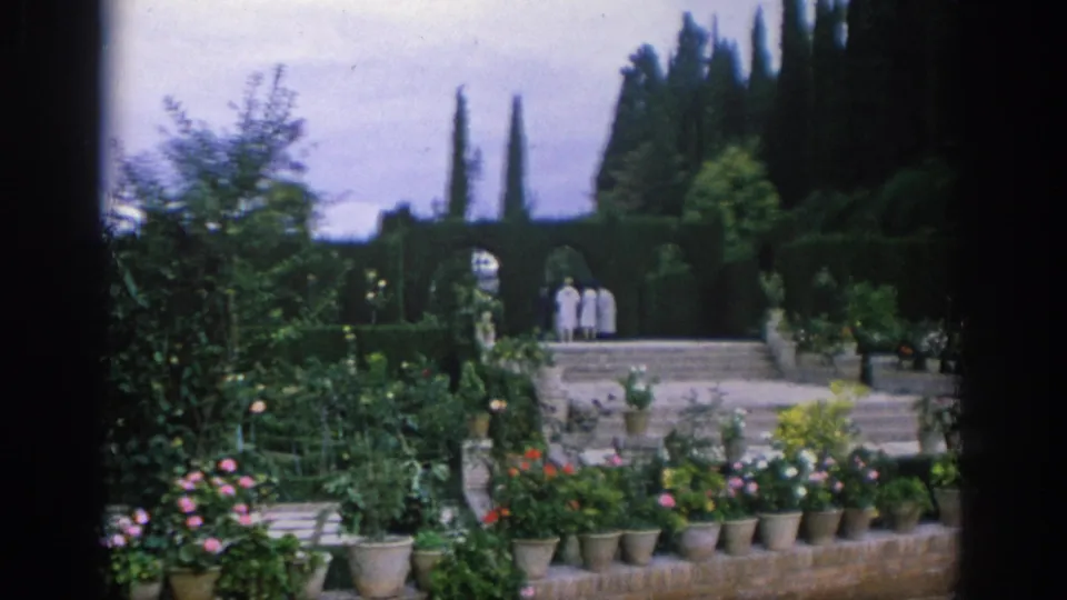 A soft 8mm view of a Spanish formal garden shows clipped hedges, flower pots, stone steps, and several distant visitors.