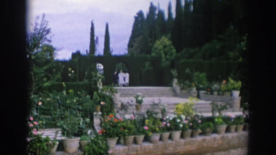 Wide 1966 Spain garden view with rows of potted flowers, clipped hedges, stone steps, tall trees, and two visitors near the upper path.