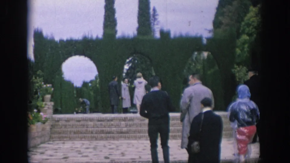 Visitors in period clothing walk across stone steps in a formal Spanish garden with hedge arches and tall cypress-like trees.