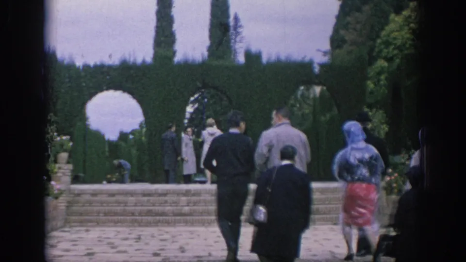 1966 Spain home-movie frame of tourists approaching brick steps, clipped hedges, and garden arches.