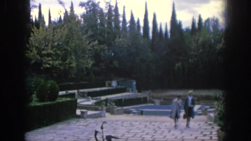 Two visitors cross a paved courtyard in a formal garden with low pools, hedges, and cypress trees in Spain.