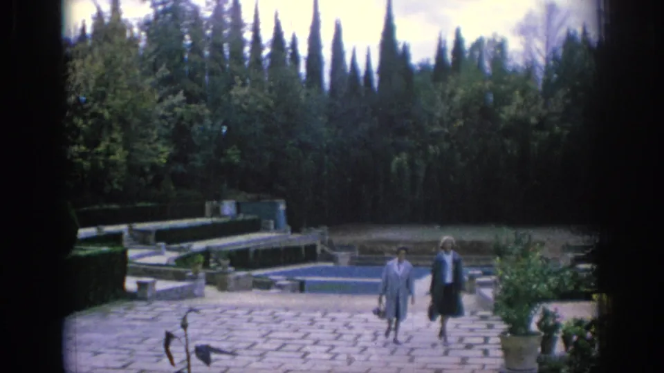 1966 Spain frame showing two women in coats crossing a paved formal garden area with pools and tall trees.