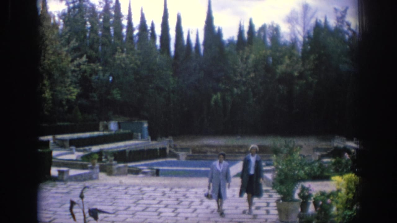 Two women walk across a stone courtyard beside a rectangular pool in a garden.