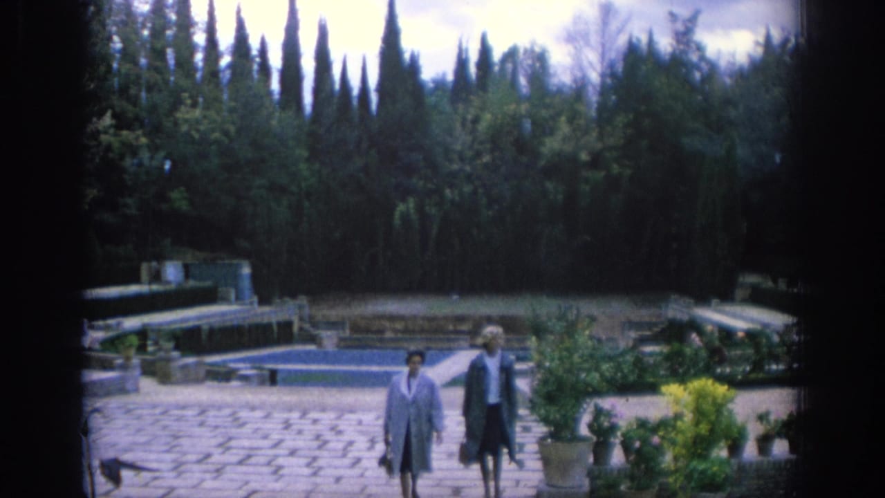 In Spain, 1966, two women approach through a stone-paved formal garden with low pools, flower beds, hedges, and cypress trees.