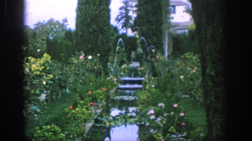 A 1966 Spain garden view shows arcing fountains over narrow pools, with flower beds, hedges, cypress trees, and a pale building partly visible.