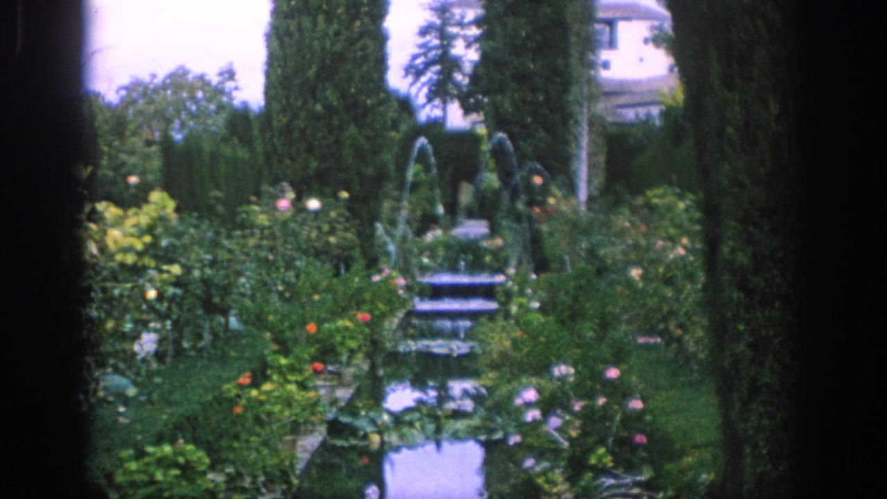 1966 Spain 8mm frame showing a formal garden with a fountain, flowering beds, tall cypress trees, and a pale building in the background.