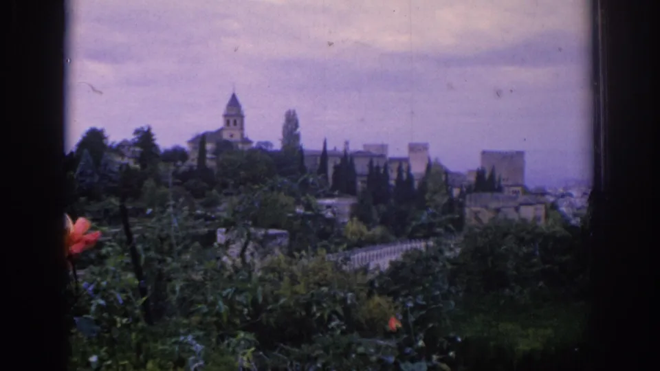 1966 Spain 8mm view toward an old town, with greenery in the foreground, stone architecture, and a church-like tower under cloudy sky.