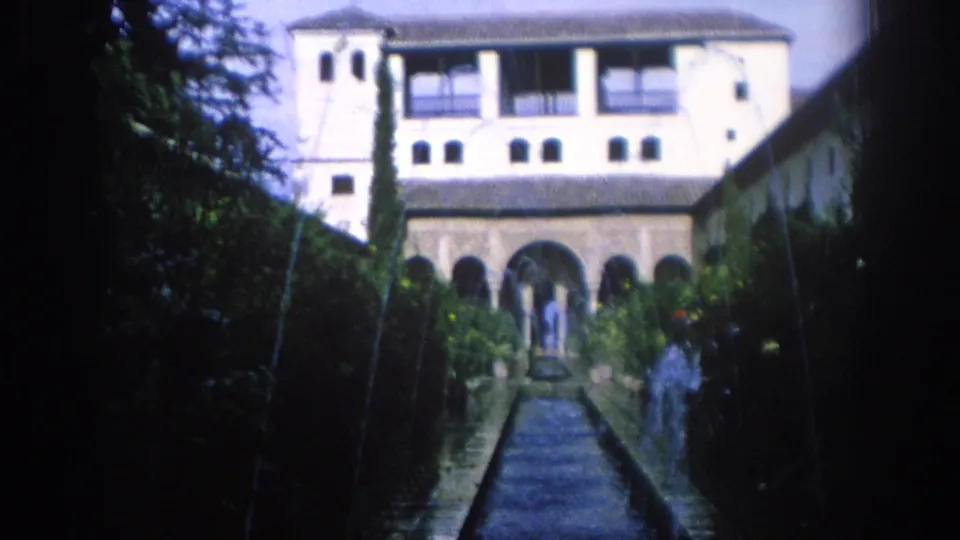 1966 Spain 8mm frame centered on a formal courtyard with a long pool, arched arcade, balcony level, hedges, and distant people.