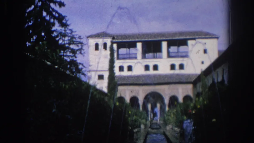 1966 Spain 8mm frame showing a pale Moorish-style courtyard facade, arched lower level, balconies, cypress tree, and distant visitors.