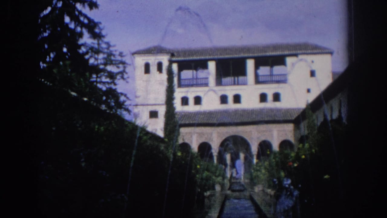 1966 Spain 8mm frame of a white two-story courtyard building with arched lower level, tile roof, garden path, and trees.