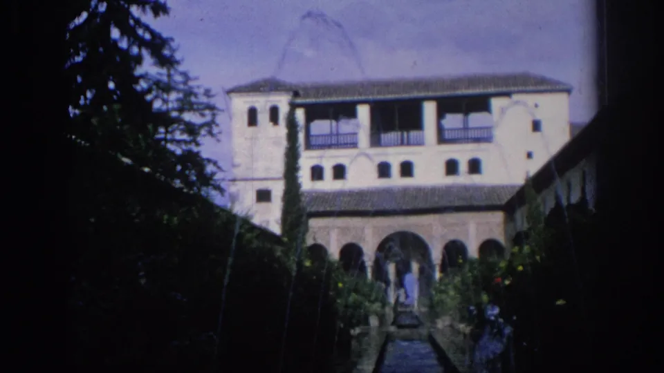 1966 Spain 8mm frame of a white two-story courtyard building with arched lower level, tile roof, garden path, and trees.