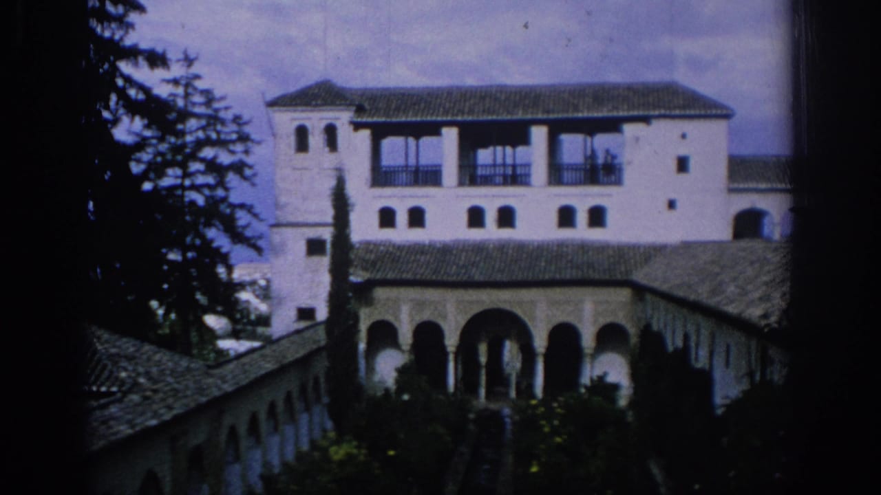 1966 Spain home-movie frame showing a white courtyard facade, arched arcade, tile roofs, cypress-like tree, and garden plantings.