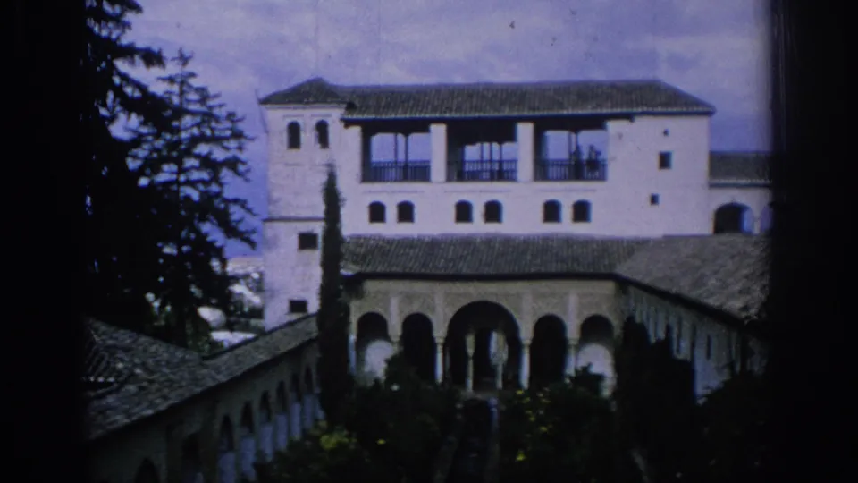 1966 Spain home-movie frame showing a white courtyard facade, arched arcade, tile roofs, cypress-like tree, and garden plantings.