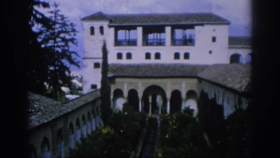 1966 Spain 8mm frame with a historic-looking courtyard garden, white arched building, tiled roofs, tall trees, and dense planting.