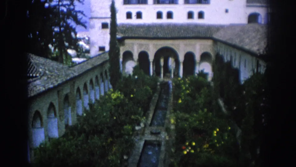 1966 Spain frame of an arched courtyard building, central water channel, planted garden beds, tiled roofs, and distant visitors.
