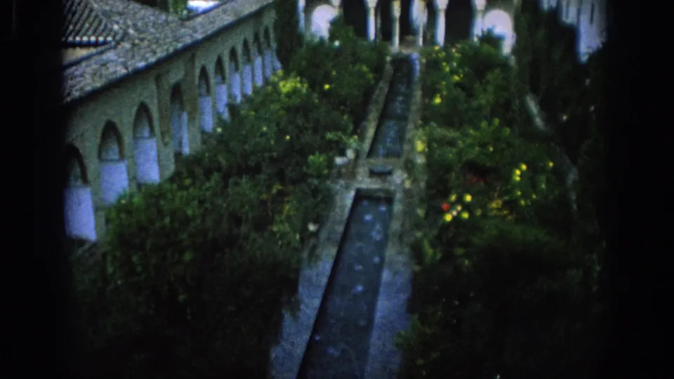 1966 Spain home-movie still with a formal courtyard garden, arched walkway, tile roof, and straight water channel viewed from above.