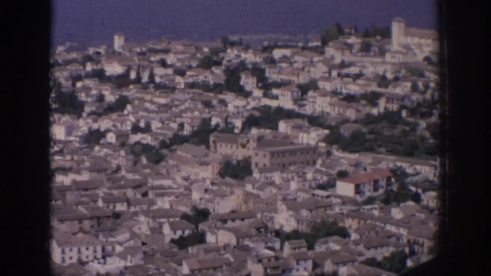 1966 Spain frame with clustered rooftops, church-like towers, trees, and a prominent ridge-top building above the city.