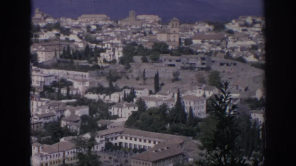 1966 Spain frame showing a hillside city district with a large masonry complex, dark trees, rooftops, and distant tower forms.