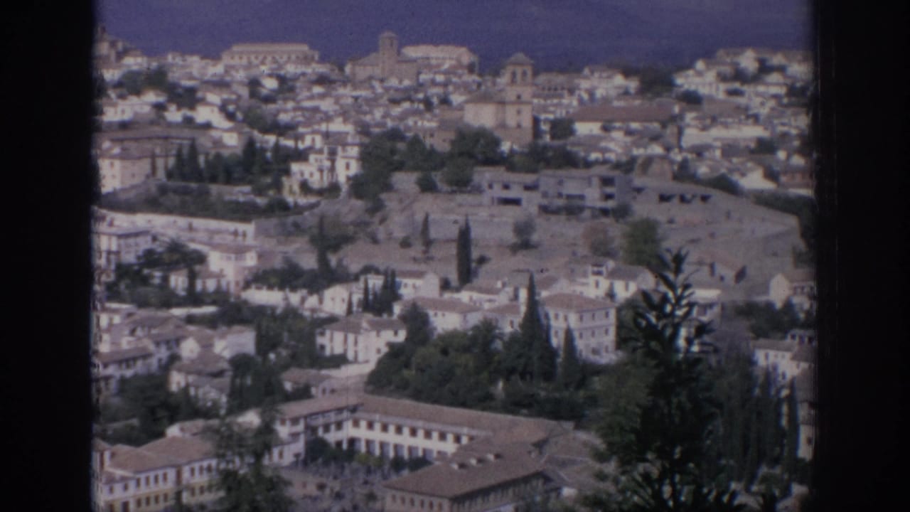 Elevated view of a hillside town with pale buildings, tiled roofs, trees, and dark film borders.