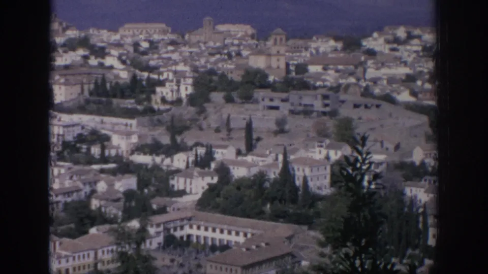 1966 Spain home-movie frame showing a hillside town from above, with white buildings, terracotta roofs, cypress trees, and soft 8mm color.