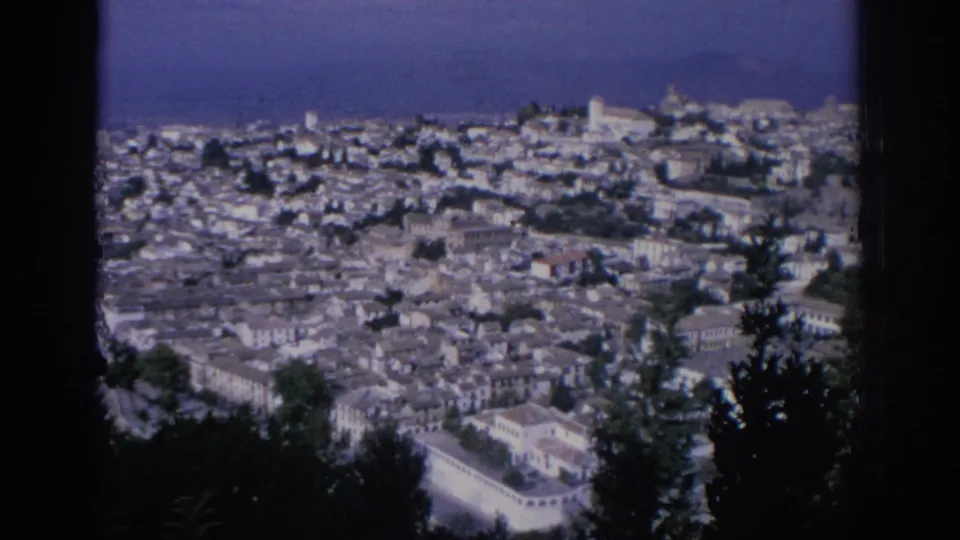 1966 Spain home-movie view across a dense hillside town, with foreground cypress silhouettes, pale rooftops, and distant slopes.