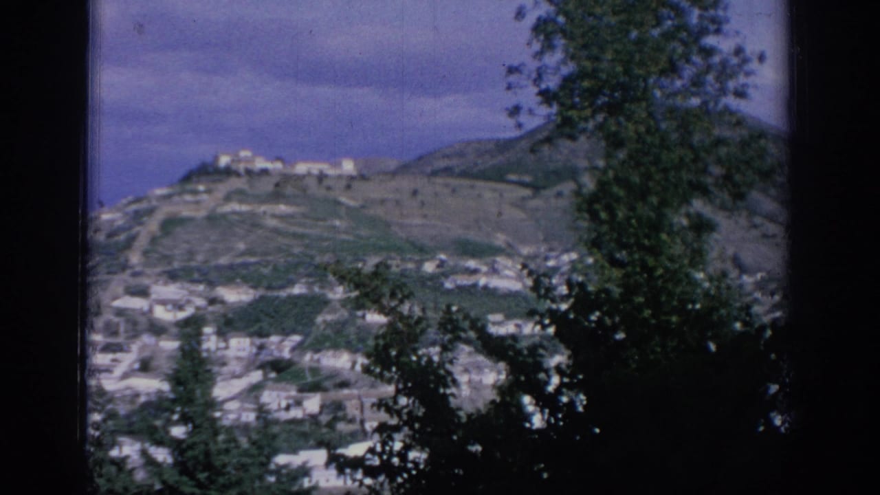 1966 Spain 8mm frame showing a distant mountain town, white buildings on slopes, trees in silhouette, and a hilltop structure.