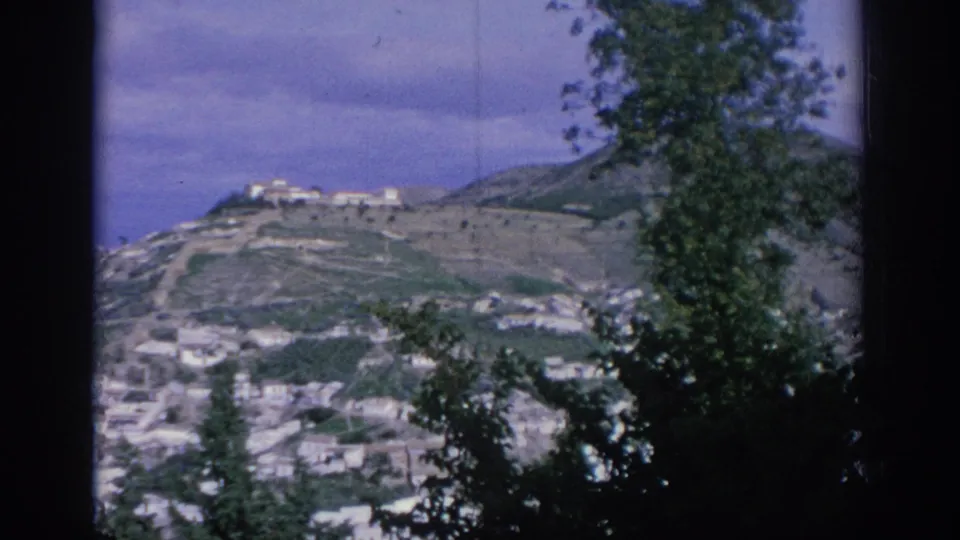 1966 Spain home-movie view of white buildings across a mountain slope, with a hilltop structure and leafy trees framing the scene.