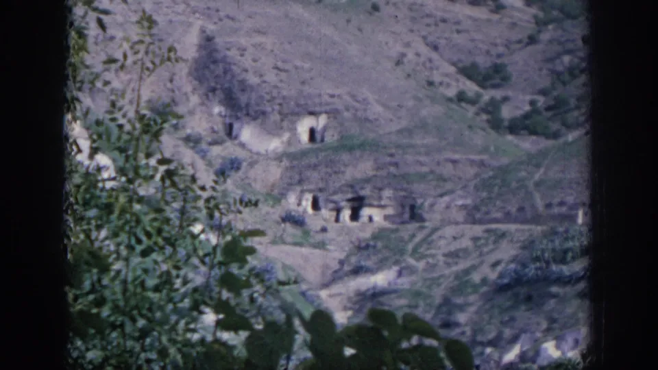 1966 Spain 8mm frame showing cave-like dwellings cut into a dry hillside, with paths, sparse vegetation, and leaves in the foreground.