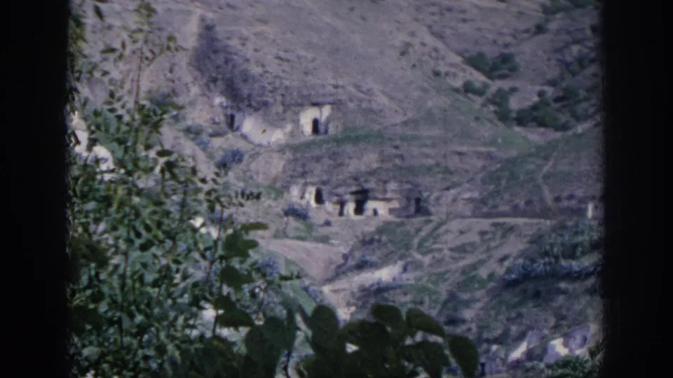 1966 Spain travel footage frame showing hillside cave dwellings, pale earth slopes, footpaths, and foreground foliage.