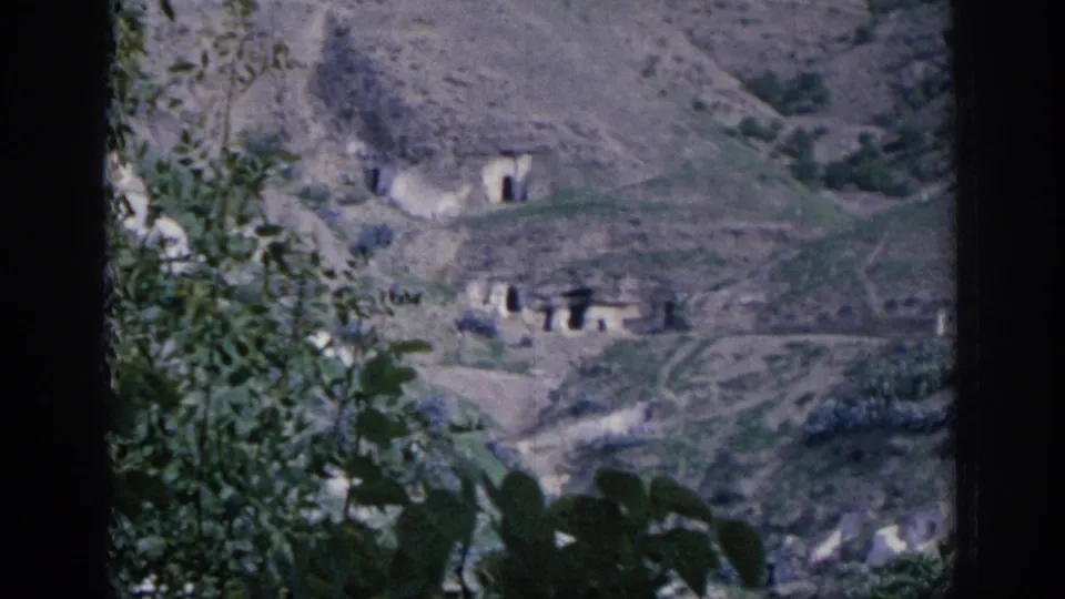 1966 Spain 8mm frame showing cave-like openings cut into a hillside, with vegetation and rugged paths visible from a distance.