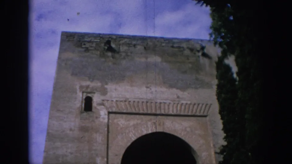 1966 Spain 8mm frame of a historic-looking stone gateway with an arched entrance and weathered masonry, partly framed by trees.