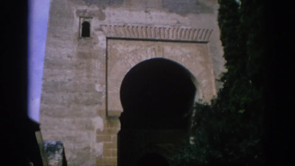 1966 Spain 8mm frame of a weathered stone archway with a dark central opening and trees partly obscuring the side.