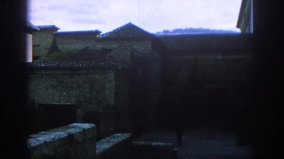 1966 Spain travel frame looking from shade toward roof tiles, old stone walls, a vertical column, and a small distant person.