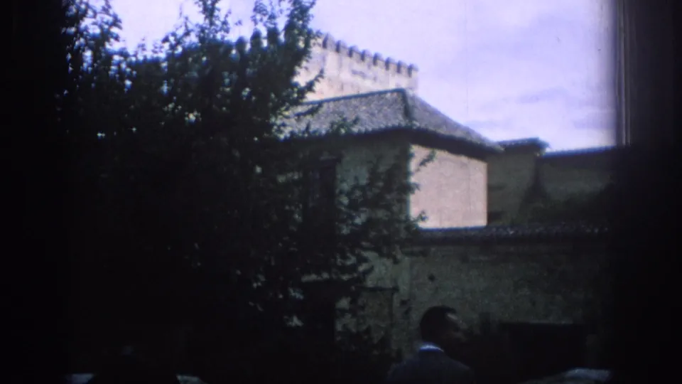 1966 Spain travel frame with a leafy tree, pale historic walls, tiled roofs, and a partial visitor in the foreground.