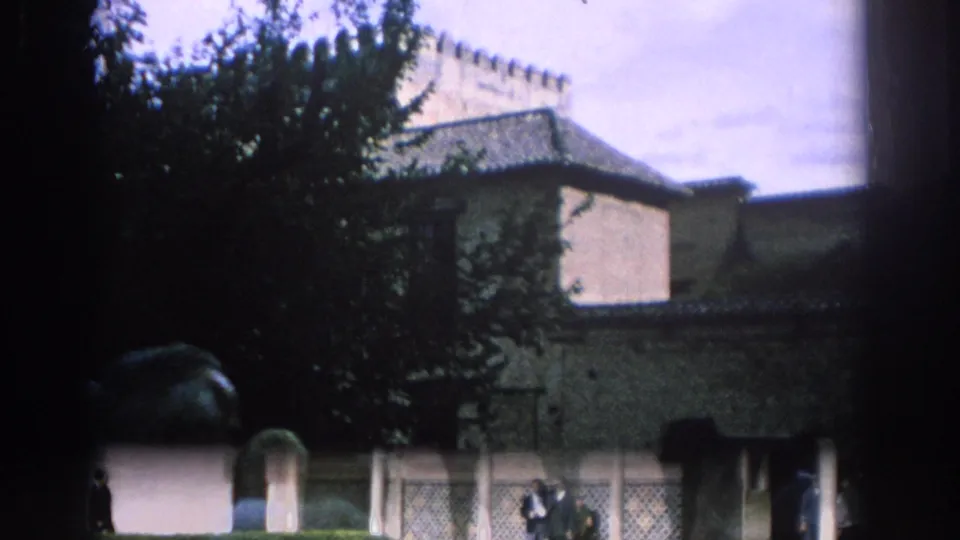 1966 Spain 8mm view of a garden edge, historic walls, tiled roofs, and visitors moving near an entrance area.