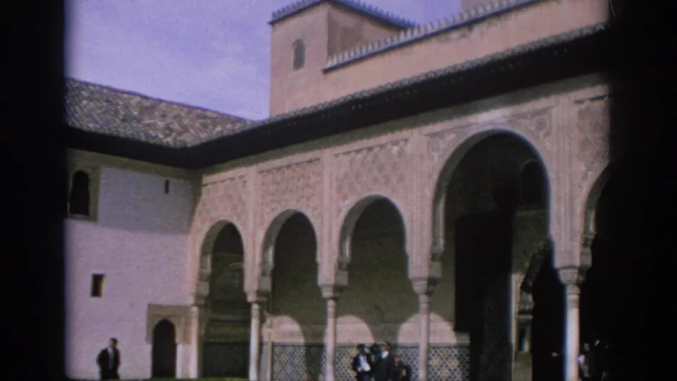 1966 Spain frame showing a sunlit Moorish-style courtyard with ornate arches, carved wall panels, visitors, and a central fountain.