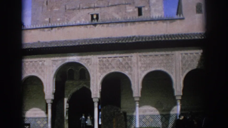 1966 Spain home-movie frame showing carved courtyard arches and columns, with a few distant visitors partly visible in the shaded arcade.