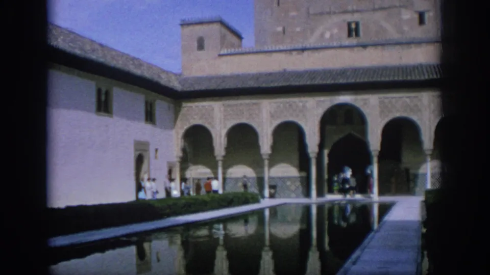 Wide 1966 Spain travel frame showing a historic courtyard, long reflecting pool, arched facade, white side wall, and visitors near the arcade.