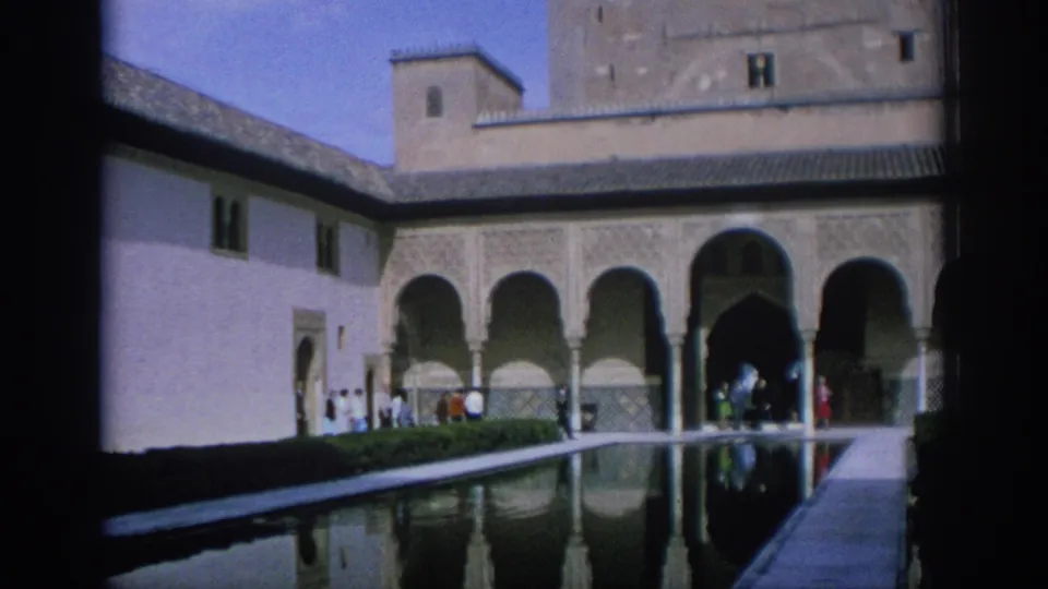 1966 Spain home-movie frame showing a long reflection pool, Moorish-style arches, and tourists moving through a historic courtyard.