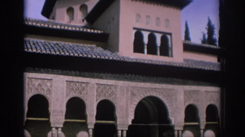 1966 Spain travel frame with ornate arched colonnades, patterned wall panels, tiled roof, upper tower windows, and trees behind the structure.