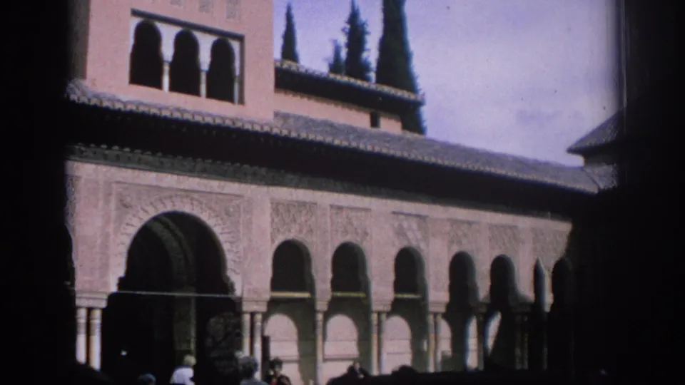 1966 Spain 8mm frame showing a historic courtyard facade with a large arch, smaller arched bays, tiled roof, cypress trees, and distant visitors.