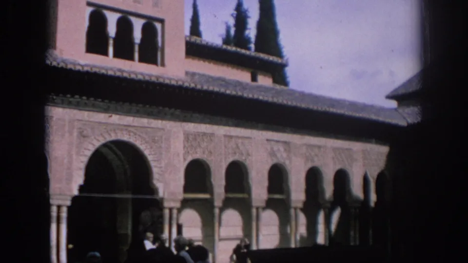 1966 Spain travel footage frame with Moorish-style arches, tiled rooflines, upper windows, cypress trees, and silhouetted tourists near the base.