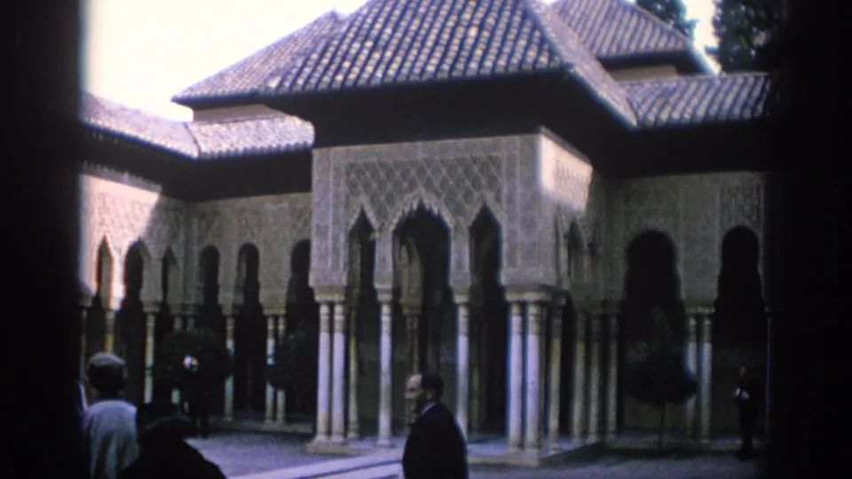 1966 Spain frame showing tourists in period clothing crossing a courtyard before ornate arches, tiled roofs, and columned walkways.
